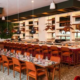 Stylish urban restaurant interior with long communal tables and orange leather chairs, marble-topped bar with red tile front and backlit glassware shelves, hanging black and palm-shaped pendant lights, and a living greenery wall.