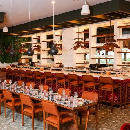Stylish urban restaurant interior with long communal tables and orange leather chairs, marble-topped bar with red tile front and backlit glassware shelves, hanging black and palm-shaped pendant lights, and a living greenery wall.
