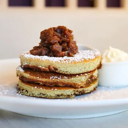 Stack of three fluffy pancakes on a white plate, dusted with powdered sugar and topped with caramelized apple compote, served with a dollop of whipped cream — sweet brunch breakfast.