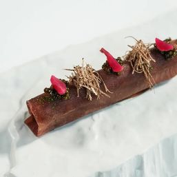 Long dark chocolate tube dessert on a white marble plate, topped with green pearl-like spheres, bright pink edible petals and golden crispy shreds — elegant fine-dining plated dessert.