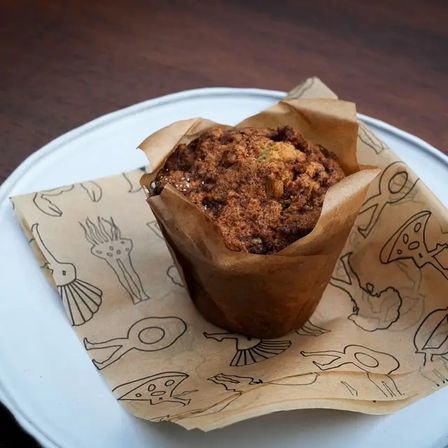Crumb-topped artisan muffin in a brown tulip paper liner on illustrated parchment, served on a white plate atop a wooden table — bakery-style breakfast pastry.