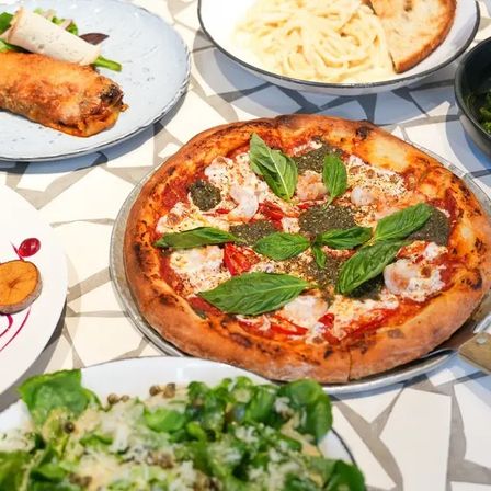 Mouthwatering Italian-style pizza topped with pesto, fresh basil and roasted peppers, served with pasta, green salad and appetizers on a patterned table