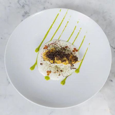 Top-down view of a gourmet plated dish on a white plate over marble: a yellow quenelle topped with dark shavings, nestled in white foam with tiny edible flowers and vivid green sauce streaks — elegant fine-dining presentation.