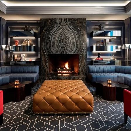 Cozy modern lounge with dramatic veined marble fireplace, tufted tan leather ottoman, curved blue sectional sofas, geometric rug, built-in lit shelves and warm ambient lighting.