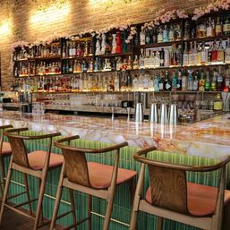 Stylish urban cocktail bar interior with a polished marble countertop, green tiled bar front and wooden stools with pink cushions, backlit shelves of liquor bottles on an exposed brick wall decorated with pink floral garlands.