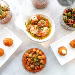 Overhead view of a Mediterranean tapas spread on a marble tabletop: garlic shrimp in olive oil, chickpea stew, marinated olives and small fried croquettes on white dishes.