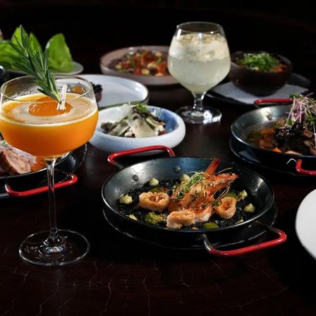 Fine-dining seafood tapas: crispy prawns and calamari in small paella pans, bright orange cocktail garnished with rosemary and assorted side dishes on a dark restaurant table.
