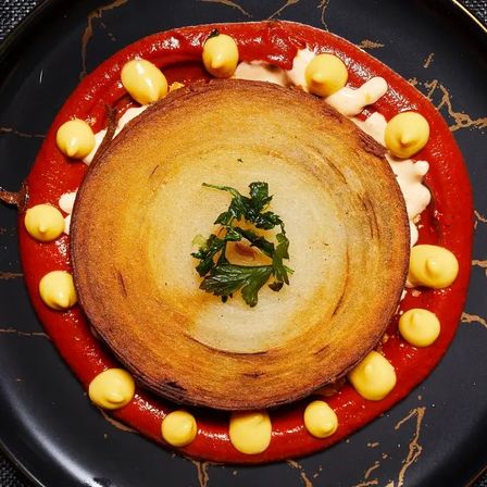 Crispy golden potato galette crowned with parsley, nestled in a ring of roasted red pepper sauce and playful yellow aioli dots on a black marbled plate — gourmet plated dish