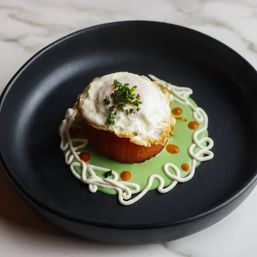 Roasted tomato cup topped with a fried egg and chopped chives, served on bright green pea purée with playful cream swirls and chili oil dots on a black plate — restaurant-style presentation.