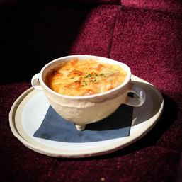 Bistro-style French onion soup with bubbling golden melted cheese and chopped parsley in a two-handled white crock, served on a plate with a dark napkin