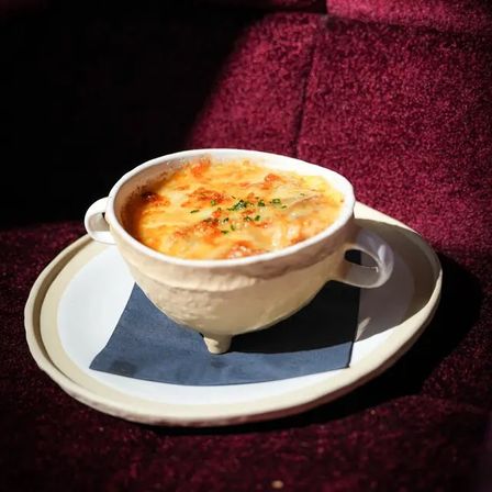 Bistro-style French onion soup with bubbling golden melted cheese and chopped parsley in a two-handled white crock, served on a plate with a dark napkin