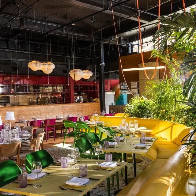 Sunlit modern restaurant interior with industrial black ceiling, yellow curved banquette, green chairs, colorful tables set with glassware, cloud-like pendant lamps and lush tropical plants.