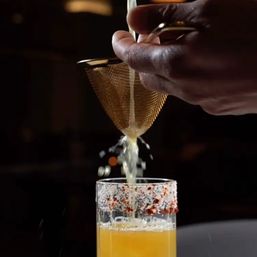 Hands straining fresh citrus through a gold mesh strainer into a chili-salt-rimmed cocktail glass, bright orange juice splashing into the drink.