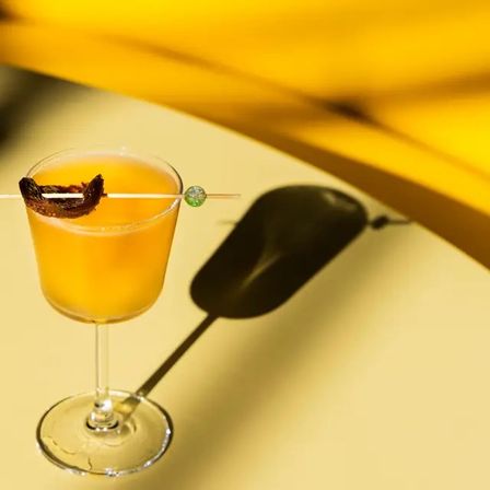 Sunlit orange cocktail in a stemmed glass garnished with dried fruit on a glass pick with a green bead, casting a playful shadow on a yellow tabletop.