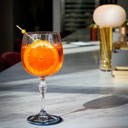Bright orange citrus spritz in a stemmed wine glass with an orange wheel and skewer, resting on a marble bar counter with a warm lamp glow in a modern cocktail lounge.