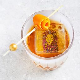 Top-down view of an amber cocktail in a patterned rocks glass with a large square ice cube featuring a colorful lion illustration and a skewered orange peel rose garnish on a light marble surface