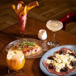 Gourmet restaurant table with two cocktails (coupe with dehydrated citrus and a frothy mango drink), a cone of plantain chips, a ceviche-style seafood salad with microgreens, and a cured meat carpaccio topped with shaved cheese, mushrooms and quail eggs.