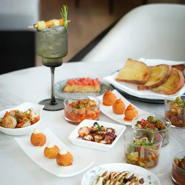 Gourmet tapas spread on a white marble table with crostini, toasted bread, marinated olives, octopus, shrimp, chickpea salad, fried croquettes, and a rosemary‑garnished cocktail