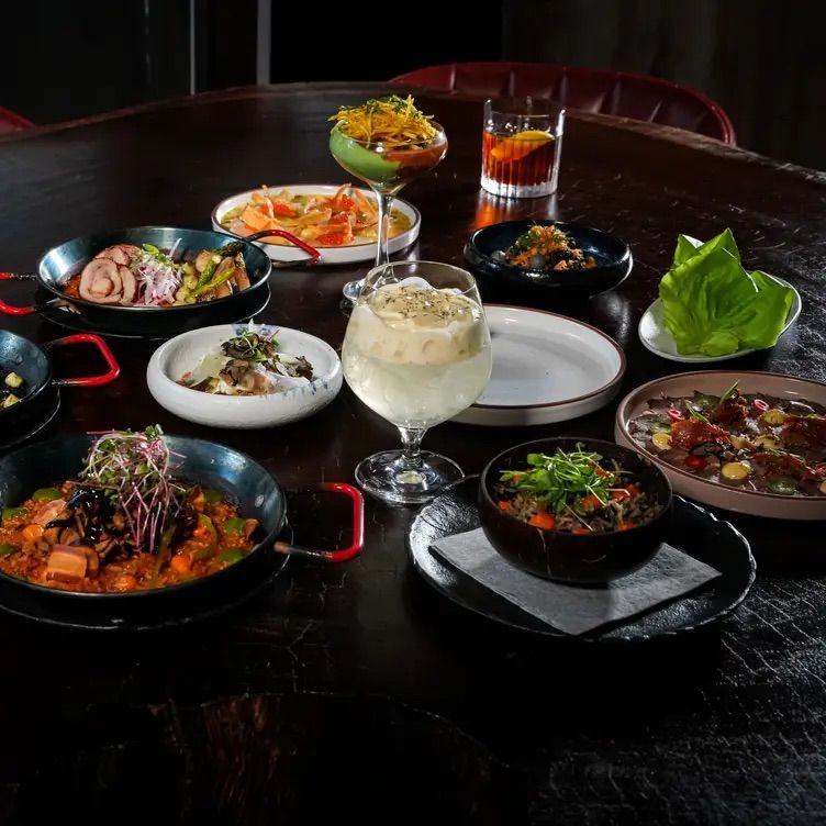 Colorful Asian-fusion small plates and bowls arranged tapas-style on a dark wooden restaurant table with fresh greens, rice and saucy dishes plus three cocktails — stylish group dining scene.