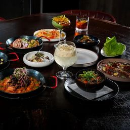 Colorful Asian-fusion small plates and bowls arranged tapas-style on a dark wooden restaurant table with fresh greens, rice and saucy dishes plus three cocktails — stylish group dining scene.