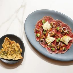 Gourmet beef carpaccio — thinly sliced raw beef topped with microgreens, pickled onion rings, mushrooms, cheese shavings and citrus zest, served with herbed crispy flatbread on a gray plate over marble.