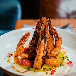 Towering grilled jumbo shrimp stacked on a creamy purée with blistered cherry tomatoes, chives, olive oil and spice drizzle — vibrant plated seafood dish at a fine-dining restaurant.