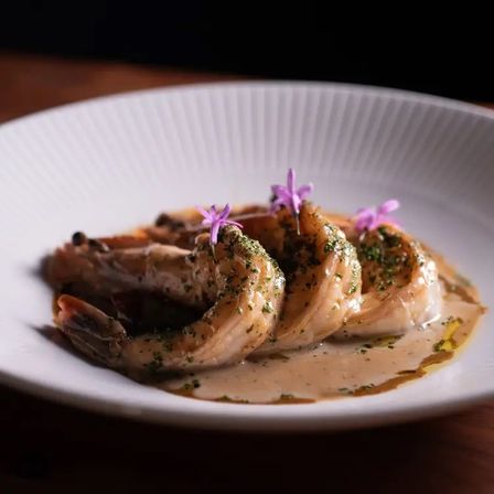 Gourmet seared prawns lounging in a creamy herb sauce, sprinkled with chopped parsley and tiny purple edible flowers on a white ridged plate