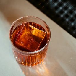 Amber cocktail in a ribbed rocks glass with a large square ice cube, sitting on a light tabletop under warm bar lighting.