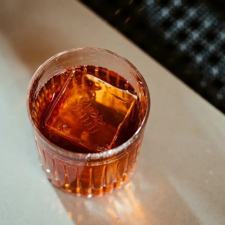 Amber cocktail in a ribbed rocks glass with a large square ice cube, sitting on a light tabletop under warm bar lighting.