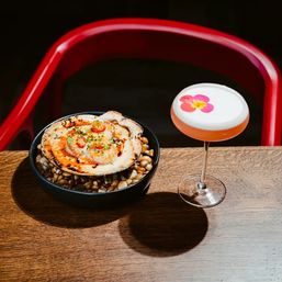 Grilled scallop in its shell on a bowl of pebbles paired with a frothy pink cocktail topped with an edible flower on a wooden restaurant table and red chair in the background.