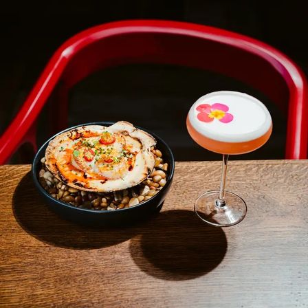 Grilled scallop in its shell on a bowl of pebbles paired with a frothy pink cocktail topped with an edible flower on a wooden restaurant table and red chair in the background.