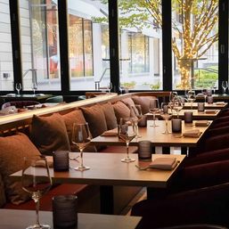 Warm, cozy restaurant interior with banquette seating and plush cushions, set wooden tables with wine glasses and candles, large windows overlooking a tree wrapped in twinkling lights on a city sidewalk.