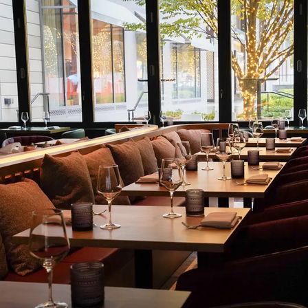 Warm, cozy restaurant interior with banquette seating and plush cushions, set wooden tables with wine glasses and candles, large windows overlooking a tree wrapped in twinkling lights on a city sidewalk.