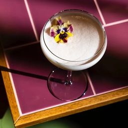 Frothy white cocktail in a coupe glass garnished with a yellow-and-purple pansy, set on glossy pink tiles with a wooden trim