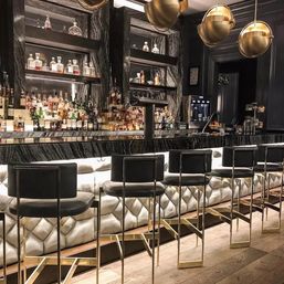 Elegant upscale cocktail bar interior with backlit quilted leather bar front, black leather and gold metal bar stools, marble back bar shelves filled with liquor bottles, and brass globe pendant lights.