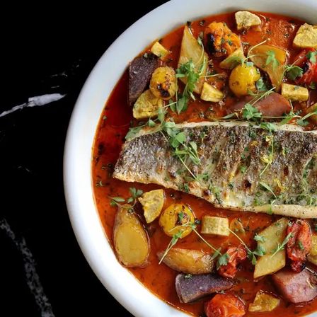 Pan-seared fish fillet with crispy skin served in a Mediterranean-style tomato broth with roasted baby potatoes, cherry tomatoes and microgreens in a white bowl.