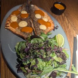 Crispy seared whole fish split open with orange and creamy sauce dots, served with mixed greens salad and lime wedge on an oval slate plate at a restaurant