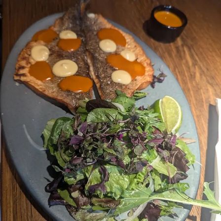 Crispy seared whole fish split open with orange and creamy sauce dots, served with mixed greens salad and lime wedge on an oval slate plate at a restaurant