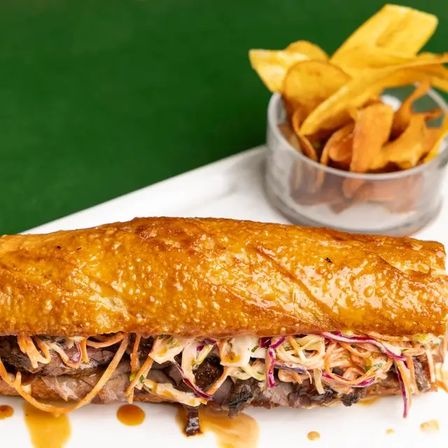 Mouthwatering golden-toasted baguette sandwich with sliced beef, creamy colorful coleslaw and savory sauce, served with crispy plantain chips on a white plate