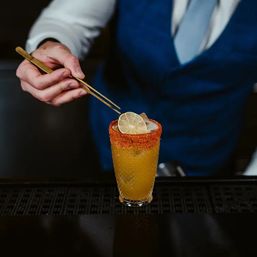 Close-up of bartender in blue vest using gold tweezers to place a lime wheel on an orange-yellow, spicy-rimmed citrus cocktail in a textured glass at a bar.