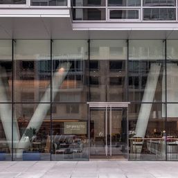 Sleek downtown glass building entrance with floor-to-ceiling windows, diagonal white support columns, double glass doors and visible seating and bar area behind the panes.
