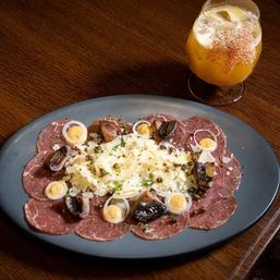 Thinly sliced beef carpaccio on a dark oval plate topped with shaved Parmesan, quail egg halves, roasted mushrooms, capers and chives, paired with an amber cocktail on a wooden table.