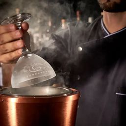 Bartender holding an etched coupe glass inverted over a copper cocktail smoker as aromatic smoke rises in a dim bar setting