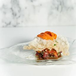 Bright uni (sea urchin) perched on a crisp rice cracker over diced tuna tartare with sesame and herbs, plated on a glass dish against a marble backdrop.