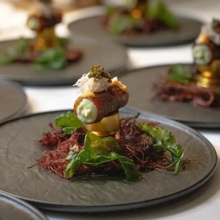 Fine-dining amuse-bouche: crispy fried tube filled with green mousse, topped with shredded seafood and green pearls, perched on a gold ring over a bed of seaweed and greens on a dark slate plate.