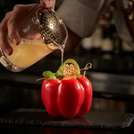Bartender strains a citrus cocktail into a hollowed red bell pepper used as a creative cup, garnished with a curled lime twist on a dark bar counter.