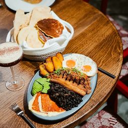 Top-down shot of a hearty Latin American breakfast on a wooden table: oval plate with sunny-side eggs, crispy pork belly and shredded beef, black beans, fried ripe plantains, sliced avocado and melted cheese arepa, plus a basket of warm arepas and a frothy cocktail.