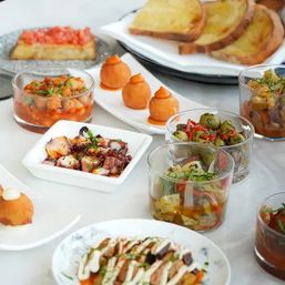 Colorful Mediterranean tapas spread on a white table: small glass bowls of marinated olives and vegetable salad, grilled octopus in a square dish, chickpea stew, crispy croquettes with aioli, bruschetta and slices of toasted bread.