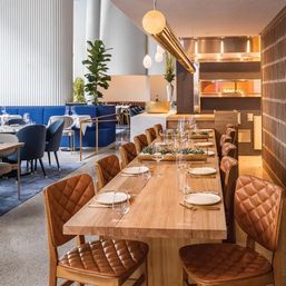 Contemporary restaurant interior with a long communal wooden table set with plates and glassware, quilted brown leather chairs, brass pendant lighting, open kitchen oven, blue banquette seating and potted greenery.