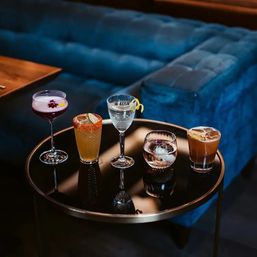 Five colorful craft cocktails — a purple coupe, a chili-rimmed citrus drink, a clear lemon-twist cocktail, a whiskey on the rocks with an ice sphere, and a brown iced cocktail — arranged on a round black glass table in a plush blue velvet cocktail lounge.